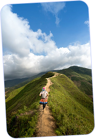 Hiker walking along a narrow dirt trail on a green mountain ridge under a blue sky with scattered clouds.